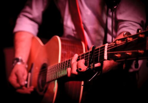 A person is playing an acoustic guitar with a capo on the neck, wearing a shirt with rolled-up sleeves, under low red lighting.