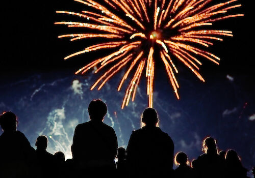 A crowd watches a large firework exploding in the night sky, silhouetted by its bright, colorful trails and glowing display above them.
