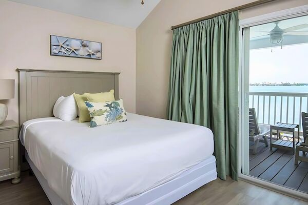 A cozy bedroom with a neatly made bed, decorative pillows, and seashell art. A sliding glass door reveals a balcony with a serene water view.