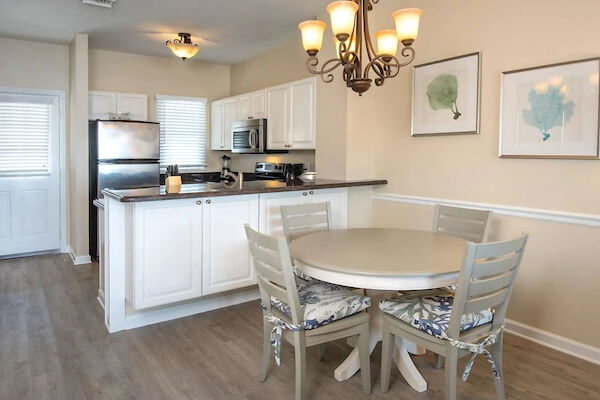 The image shows a kitchen and dining area with a round table, four chairs, a light fixture, and kitchen appliances.