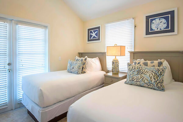 A cozy bedroom with two beds, white linens, decorative pillows, and sea-themed wall art; a lamp between the beds.