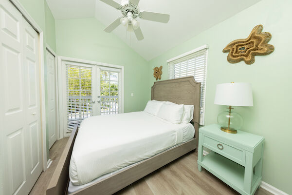 A bedroom with a bed, nightstand, lamp, closet, ceiling fan, and wall decor. Walls are light green, and there's a door with blinds.