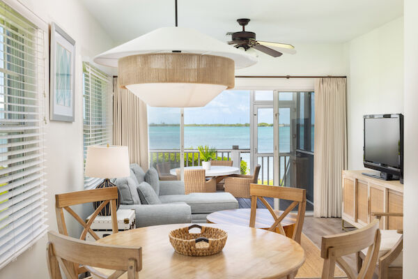 Cozy living room with wooden furniture, a gray sofa, TV, and a view of a sunny balcony overlooking a serene body of water.
