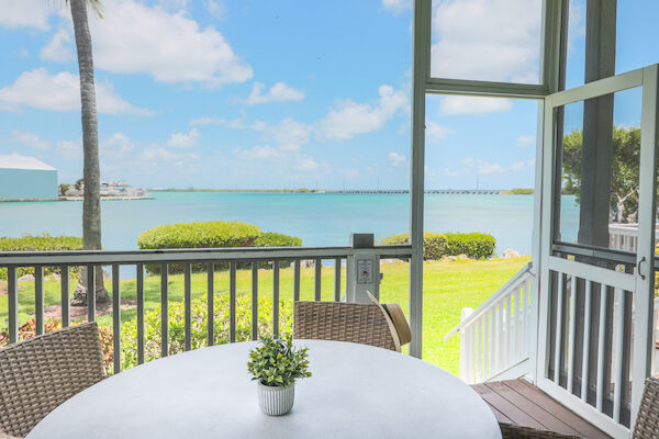 A round table with a small plant sits on a screened porch overlooking a scenic ocean view with a bridge and lush greenery.