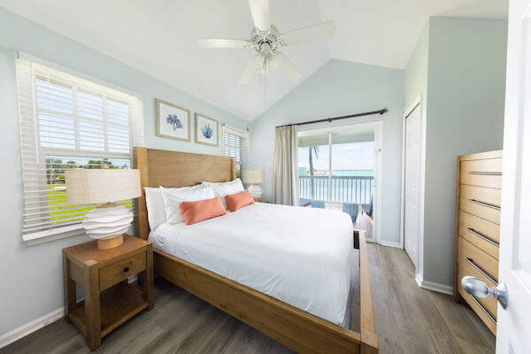 A bright bedroom with a wooden bed, white bedding, coral pillows, lamps, and ocean view through large windows and a glass door.