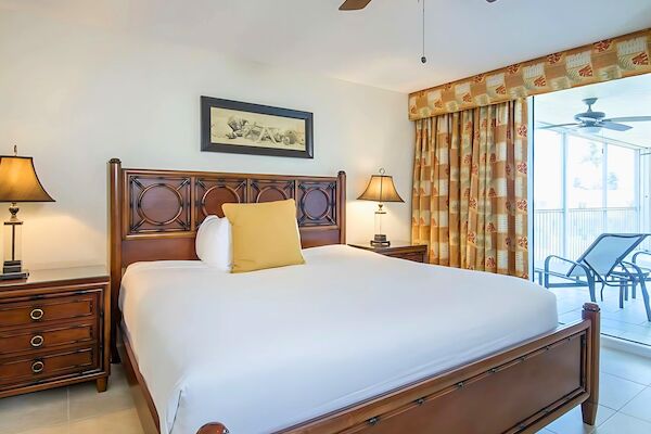 A neatly made bed with a wooden frame, bedside tables with lamps, a ceiling fan, curtains, and a balcony with chairs.
