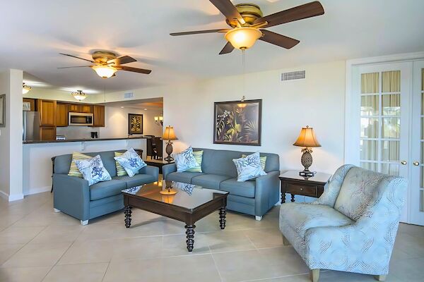 A cozy living room with blue sofas, a patterned armchair, glass coffee table, lamps, and ceiling fans; a kitchen is visible in the background.