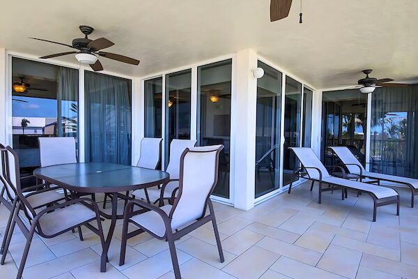 A patio features a table with chairs, sun loungers, ceiling fans, and sliding glass doors on tiled flooring.