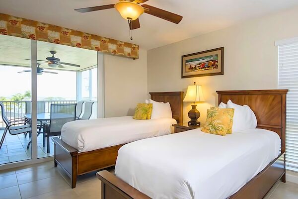 The image shows a bedroom with two twin beds, a ceiling fan, a lamp, and a painting on the wall. There's a balcony outside.