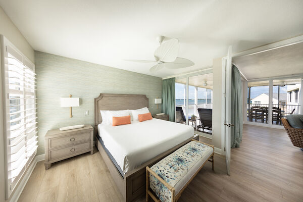 A cozy bedroom with a king-size bed, balcony view, wooden furniture, and a ceiling fan creating a bright, relaxing atmosphere.