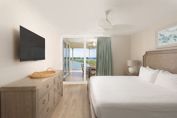 A bright bedroom with wooden furniture, a TV, a bed, artwork, and a balcony view with a table and chairs overlooking water.