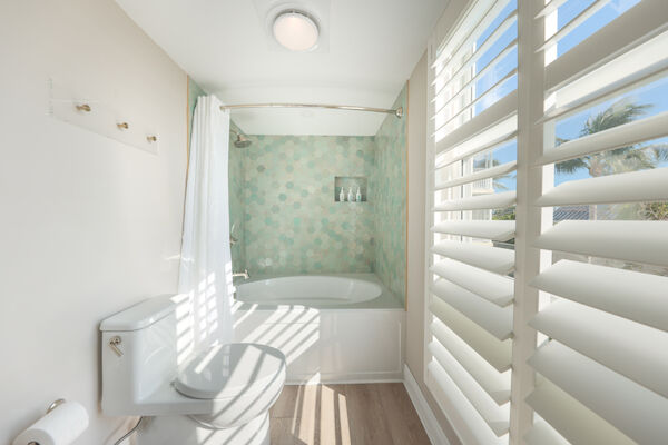 A bright bathroom with a bathtub, teal hexagonal tiles, and a white curtain. There's a toilet, wooden floor, and large window with shutters.