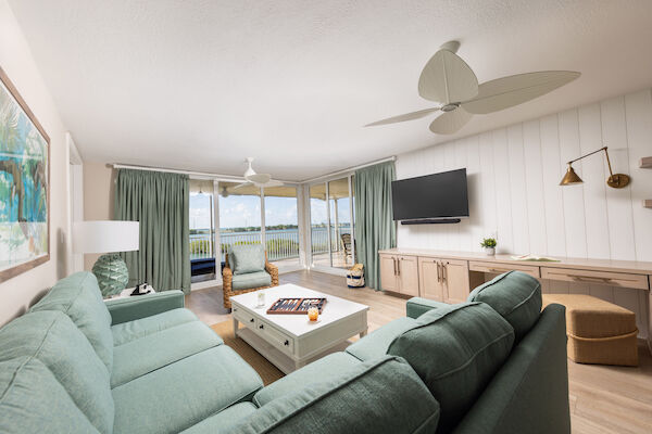A cozy living room with green couches, a wicker chair, wall-mounted TV, and large windows offering a scenic outdoor view.