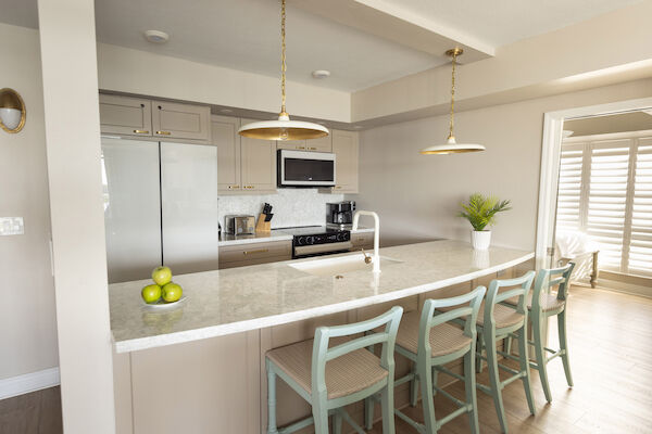 A modern kitchen with marble countertops, bar stools, hanging lights, a fruit bowl, a plant, and appliances like a microwave and coffee maker.