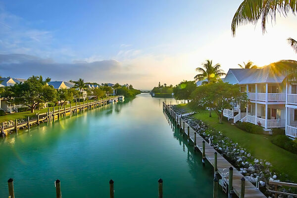 A serene waterfront scene with houses, a canal, docks, and palm trees under a clear sky at sunrise or sunset.