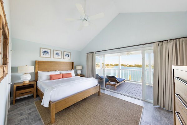 A bright bedroom with a wooden bed, coastal art, and a view of water through open sliding doors leading to a balcony with loungers.