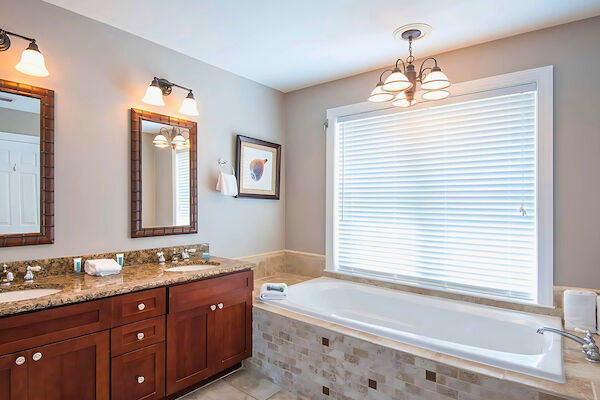 Luxurious bathroom featuring a double sink vanity with mirrors, a soaking tub, overhead lighting, and a window with blinds.