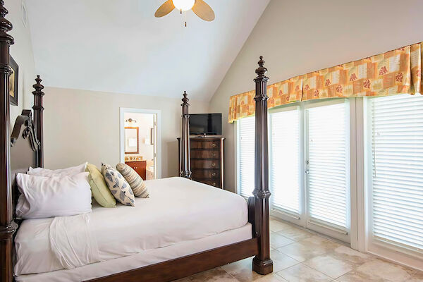 A cozy bedroom with a four-poster bed, ceiling fan, and TV. Large windows with blinds and a door visible in the background.