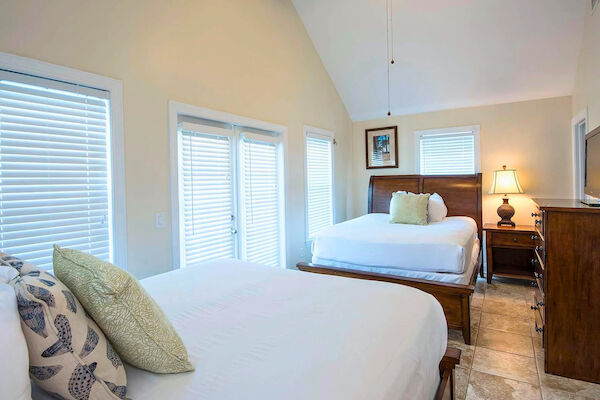 The image shows a bedroom featuring two beds, a ceiling fan, blinds on windows, a wooden dresser, and a lamp on a nightstand.