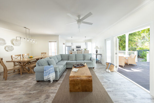 A modern living room features a sectional sofa, wooden table, open kitchen, and patio with wicker chairs, surrounded by greenery outdoors.