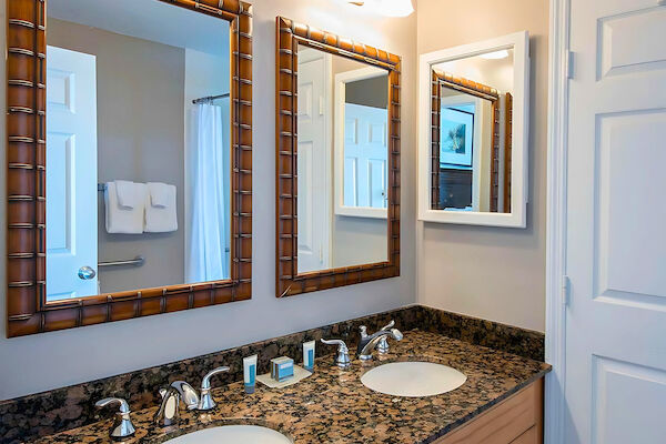 A bathroom with dual sinks on a granite countertop, two mirrors, light fixtures, and a towel bar by a shower area and door.