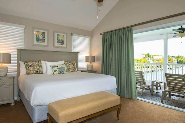 A cozy bedroom with a large bed, stylish pillows, nightstands, lamps, and art on the wall. A balcony with chairs overlooks a pleasant view.