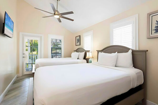 The image shows a tidy bedroom with two beds, white bedding, a ceiling fan, a small TV, and art on a pastel wall.