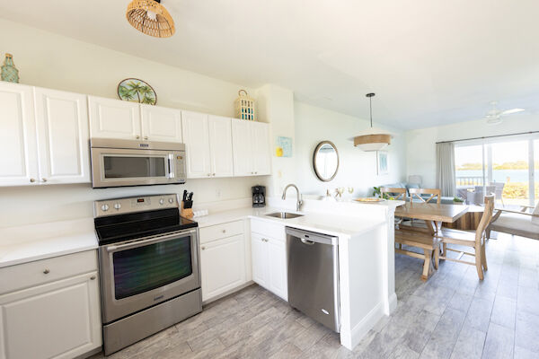 A bright kitchen with white cabinets, stainless steel appliances, and wooden dining table, overlooking a view through glass doors.