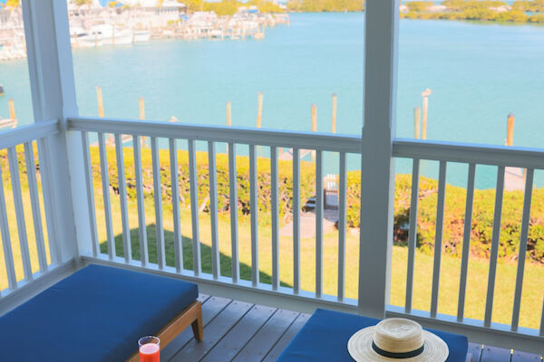 A porch with a view of the water, featuring lounge chairs, a straw hat, a magazine, a drink, and a snack on a small table.