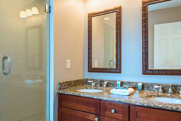 A bathroom with dual sinks, granite countertop, wooden cabinets, mirrors, light fixtures, and a glass shower enclosure.