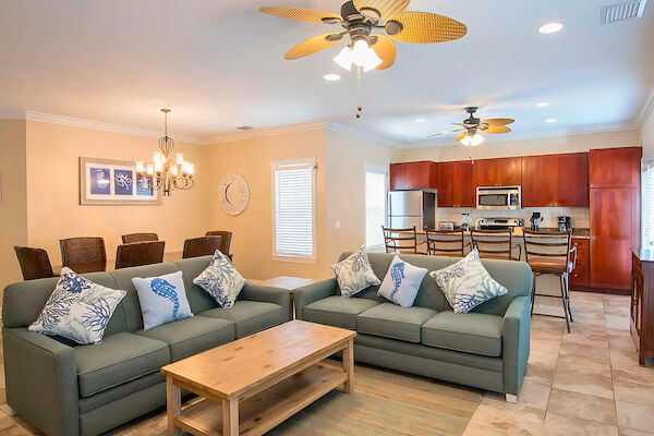 The image shows a living and dining area with two sofas, a wooden table, kitchen, ceiling fans, and decorative wall art.