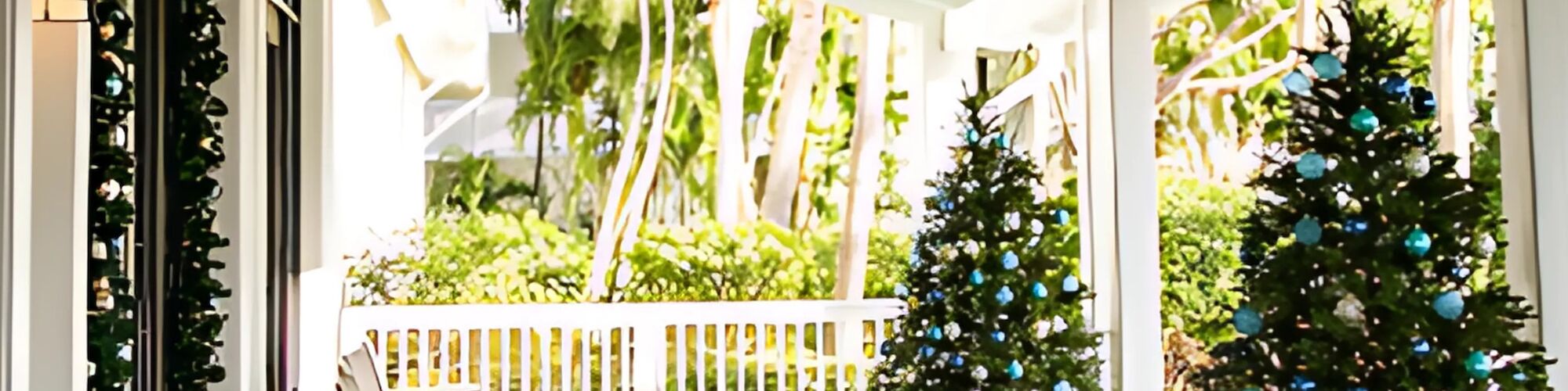 A porch with two decorated Christmas trees and rocking chairs, surrounded by greenery and a white railing.